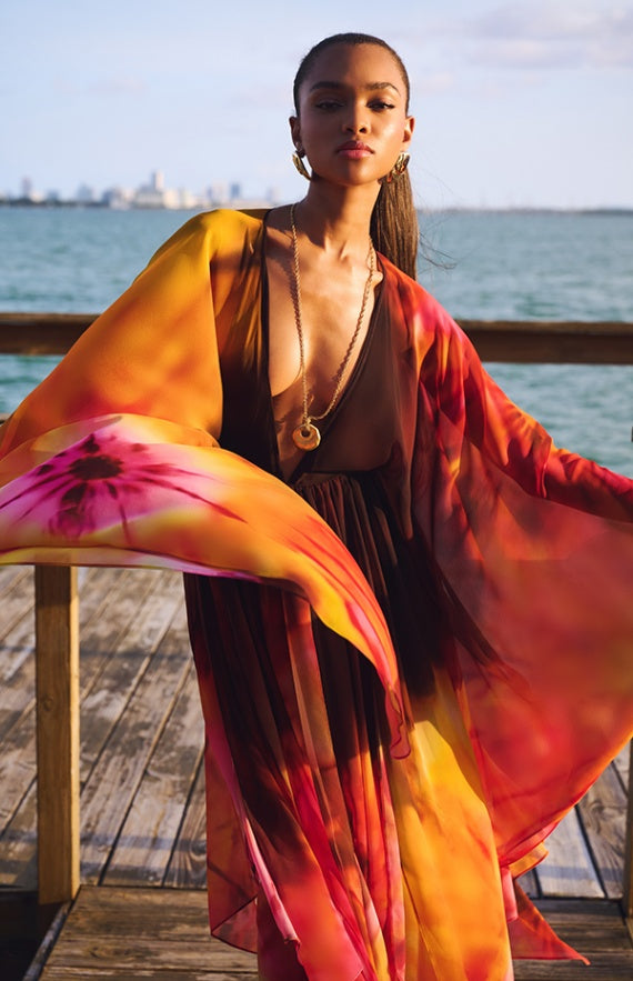 Model on a wooden boardwalk by the sea wearing a flowing multicolor tie-dye dress in yellow orange and brown with gold accessories media 1 of 4