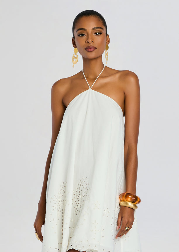 Front view of white halter neck mini dress with embroidered eyelet hem and bold gold earrings on model