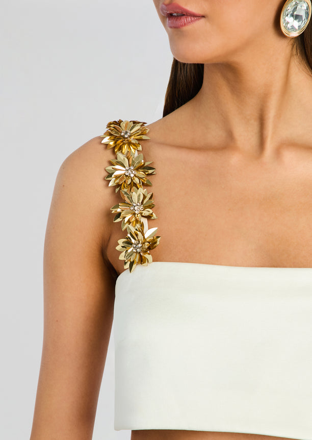 Close-up of a gold flower shoulder strap with a crystal centerpiece on a cream top.