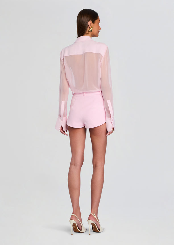 Back view of a model in pale pink sheer blouse tucked into matching shorts with belt and nude heels