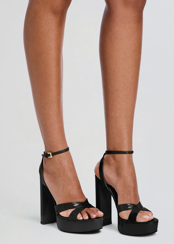 Black platform high-heel sandals with ankle straps and block heels, shown on a model's feet against a light gray studio background. media 2 of 5