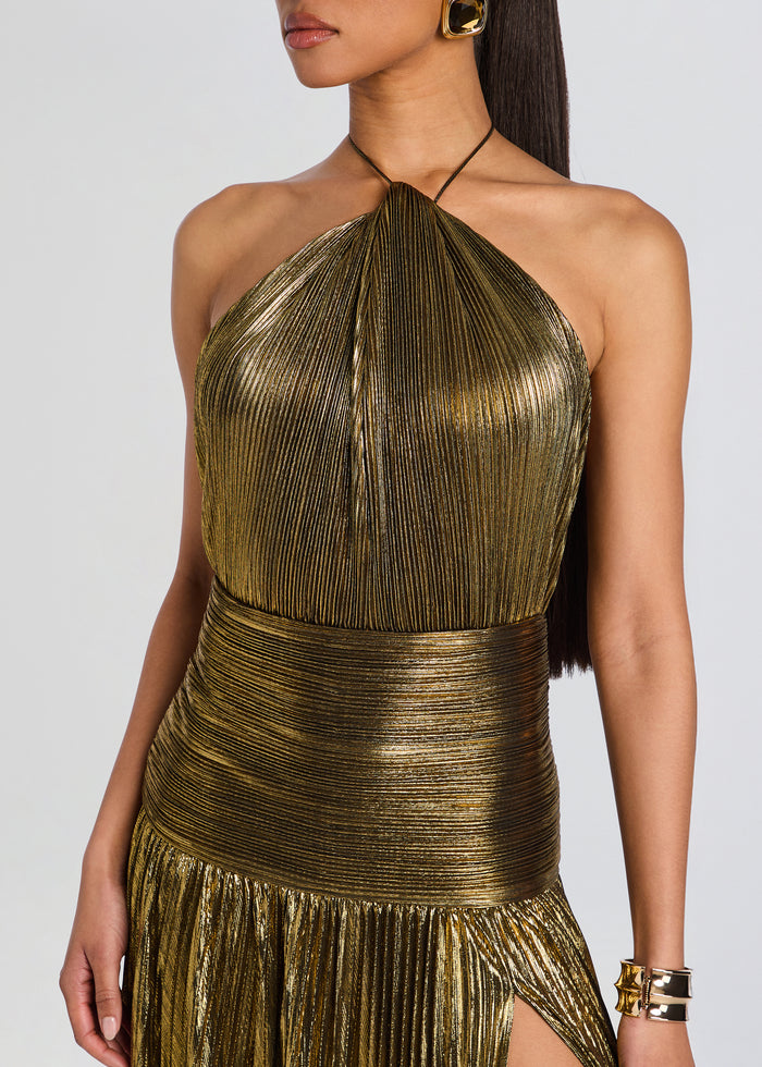 A close-up of a model wearing a metallic gold halter-neck dress with a pleated texture and a high side slit, set against a plain light gray background. media 2 of 5