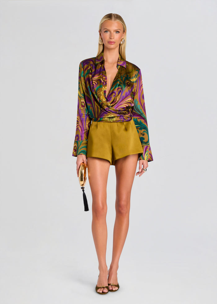 A model stands against a light gray studio background wearing a long-sleeve satin wrap blouse with a multicolor purple, green, and gold baroque-style print, paired with olive green shorts, holding a gold tassel clutch. media 2 of 5