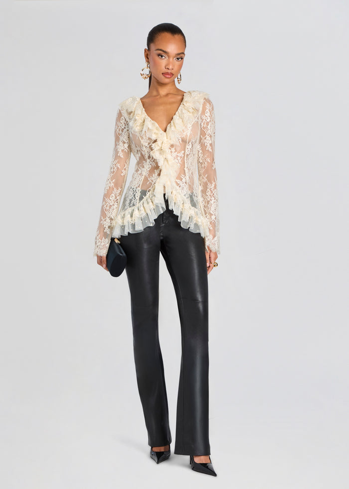 A model stands against a light gray studio background wearing a sheer cream lace long-sleeve blouse with ruffles over black leather-look pants, holding a small black clutch. media 2 of 5