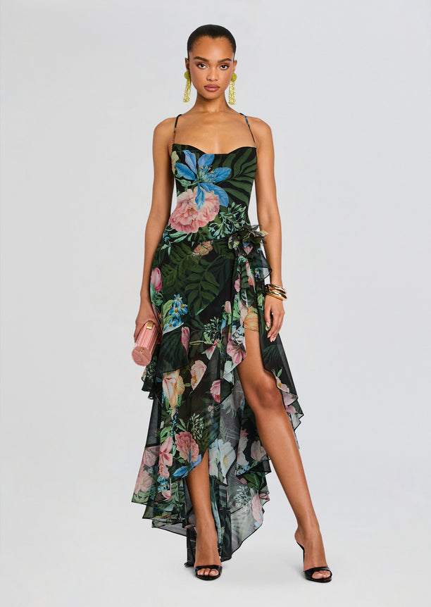 Model wearing a tropical floral silk chiffon dress with a black background and layered high low skirt, with a pink clutch and yellow earrings.