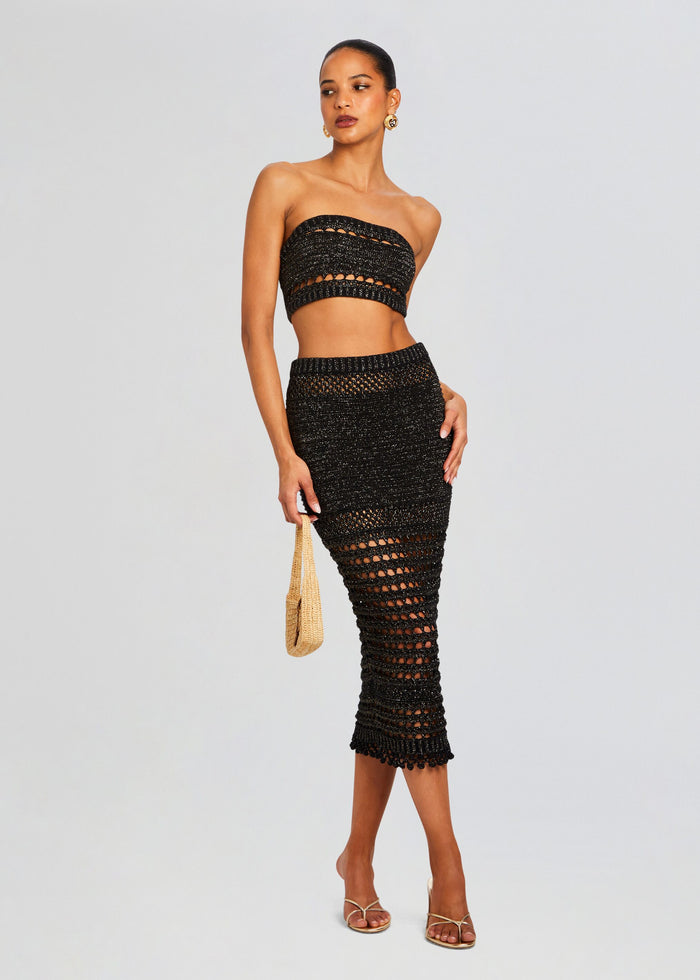Full-length model in a black Luiz Knit Crochet two-piece set with bandeau top and openwork midi skirt, holding a straw handbag. media 2 of 6