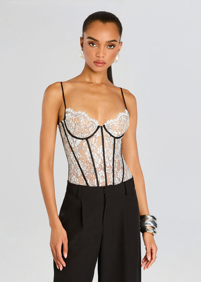 Model in a white lace corset bodysuit with black piping and thin shoulder straps, paired with black trousers. media 1 of 5