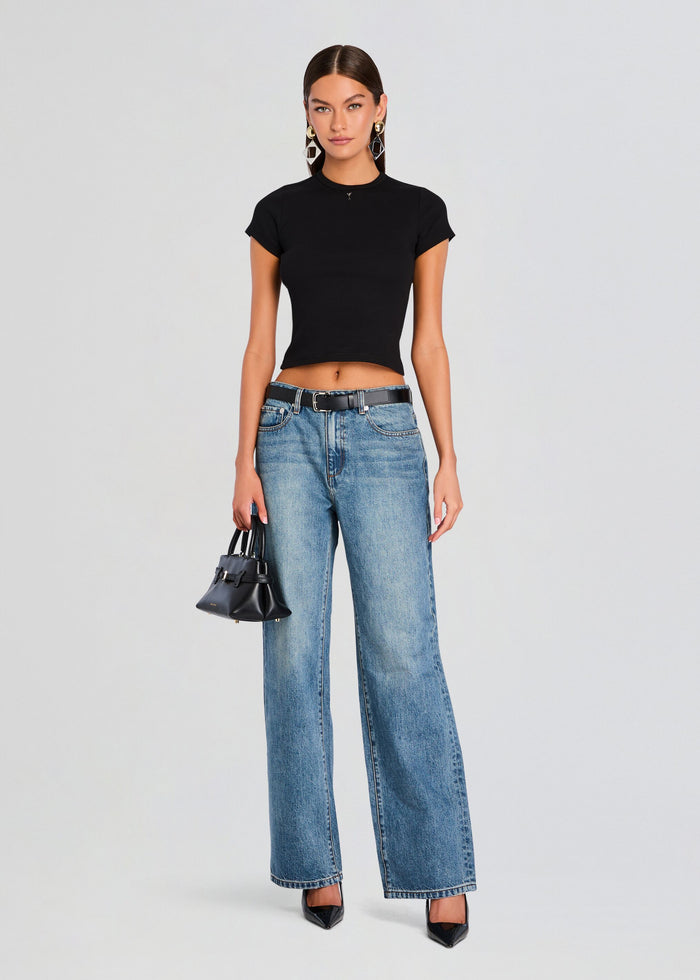Full-length shot of a model in a black cropped Tove Top with blue wide-leg jeans, black belt, and pointed-toe heels carrying a small black handbag media 2 of 5