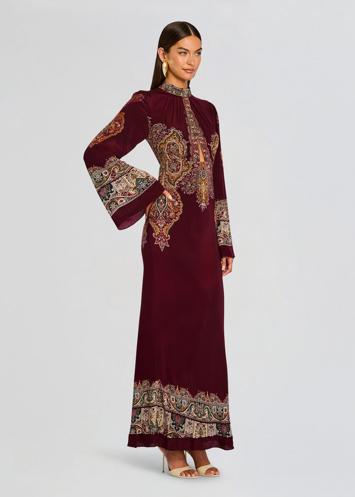Shifa Long Dress worn by a model shows a burgundy ankle length gown with dramatic patterned borders on the cuffs and hem. media 3 of 5