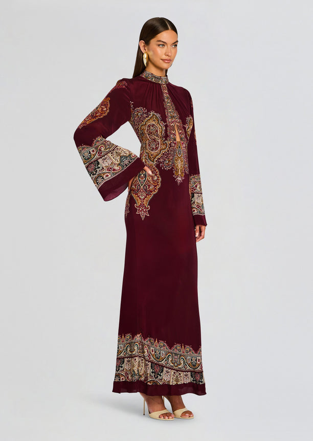 Shifa Long Dress worn by a model shows a burgundy ankle length gown with dramatic patterned borders on the cuffs and hem.