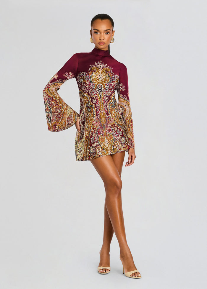 Model in a burgundy paisley print mini dress with a high neckline and flared bell sleeves, wearing gold hoop earrings. media 1 of 5