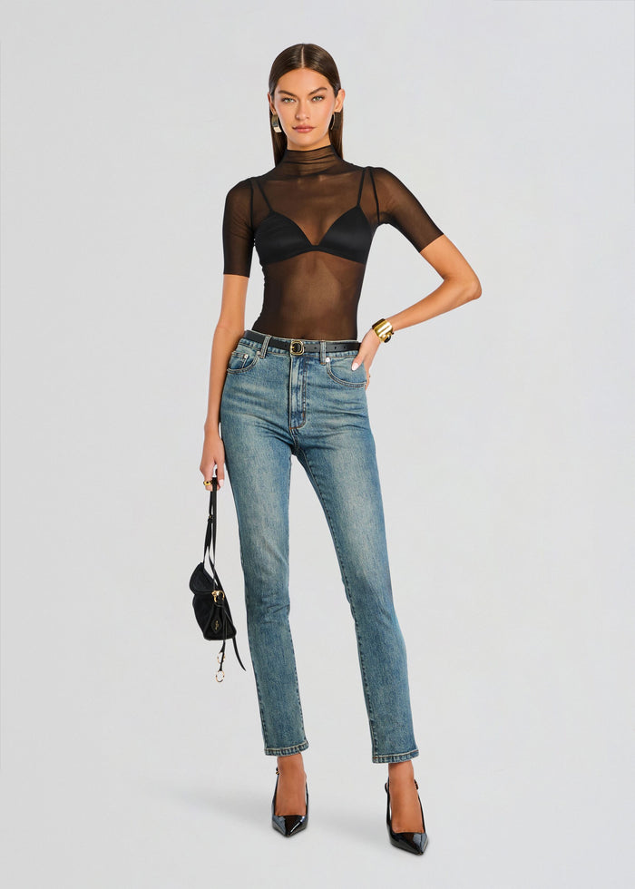 Model in a sheer black mesh short sleeve top over a black bra, paired with blue jeans and black patent heels. media 2 of 6
