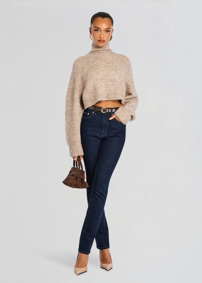 Full-length shot of a model in a beige cropped turtleneck sweater with dark jeans, a brown belt, and a small brown handbag. media 2 of 6