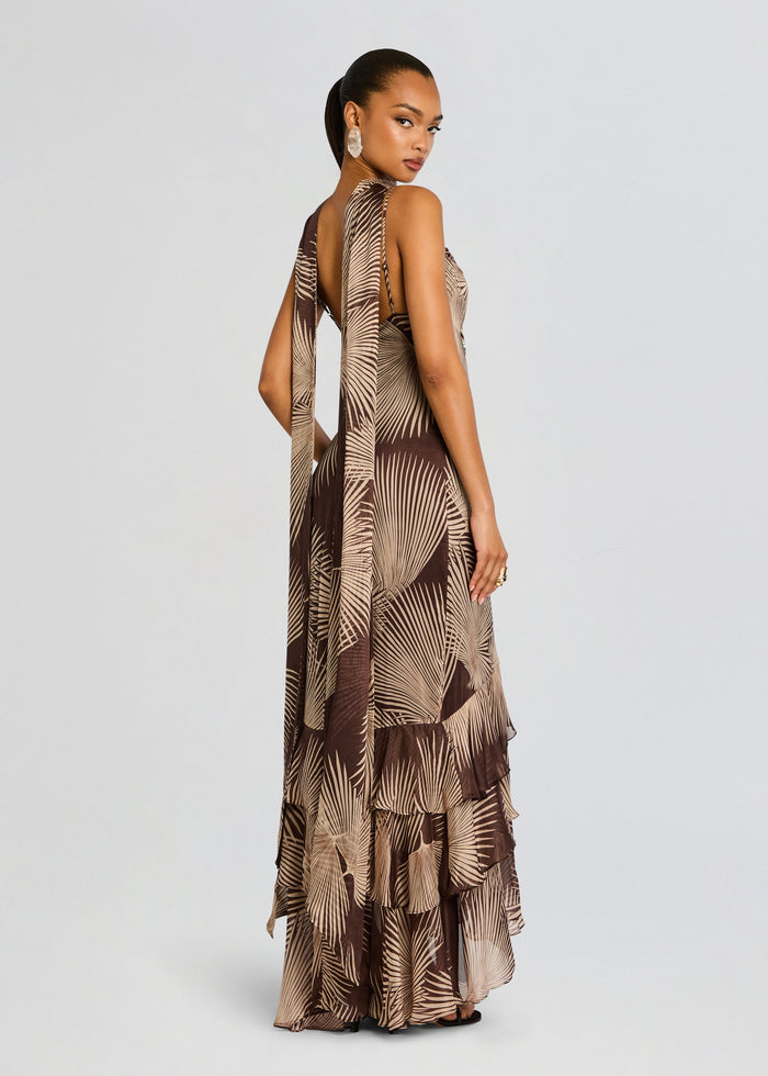 Back view of a model in a brown palm print Taro Maxi Dress with a long sash and tiered floor-length hem. media 1 of 5