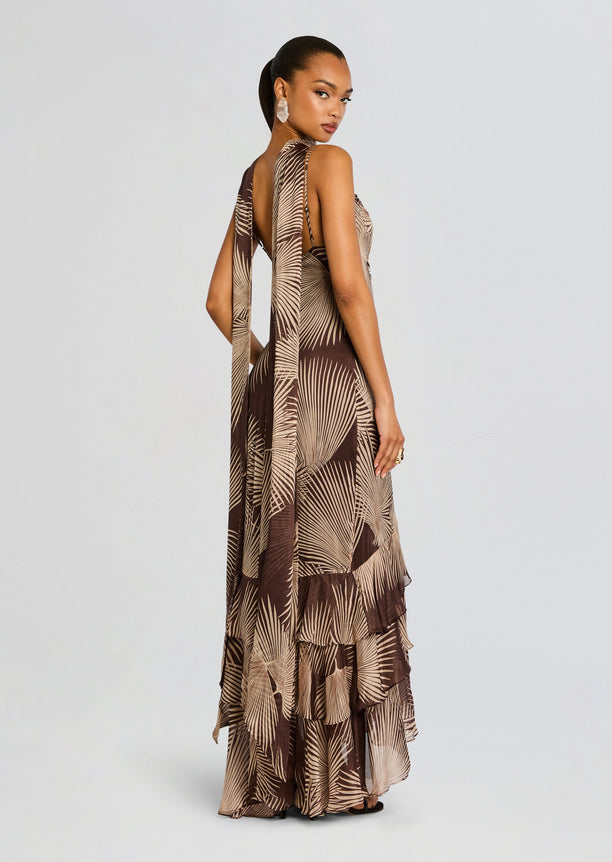 Back view of a model in a brown palm print Taro Maxi Dress with a long sash and tiered floor-length hem.