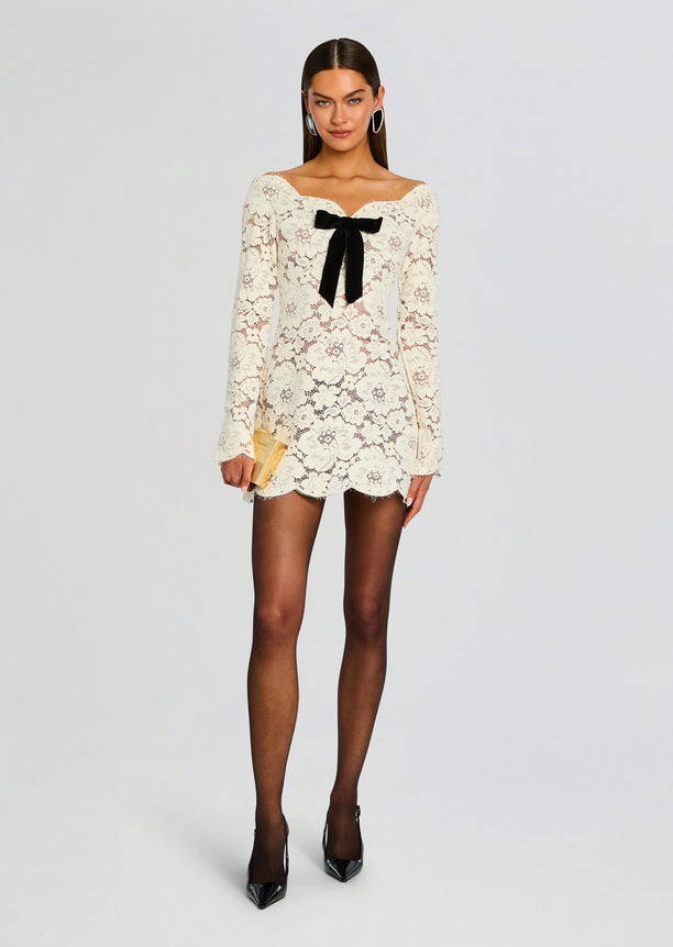 Ivory lace mini dress with long sleeves and a black velvet bow at the neckline, model holding a beige clutch.