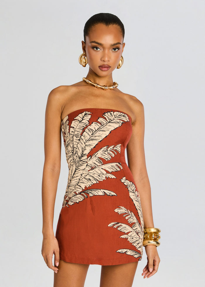 Three quarter front view of a model wearing the rust palm leaf strapless mini dress with gold jewelry. media 5 of 5