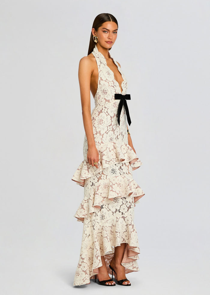 Ivory lace Senara dress with halter neckline, black satin bow at the waist, and a tiered ruffle skirt worn with black heels. media 3 of 5