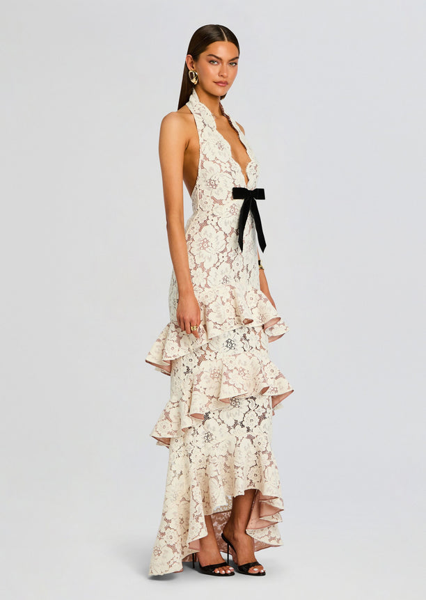 Ivory lace Senara dress with halter neckline, black satin bow at the waist, and a tiered ruffle skirt worn with black heels.