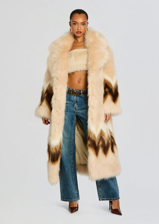 Model wearing a long beige Magali faux fur coat with brown chevron accents over a cream bandeau and jeans.