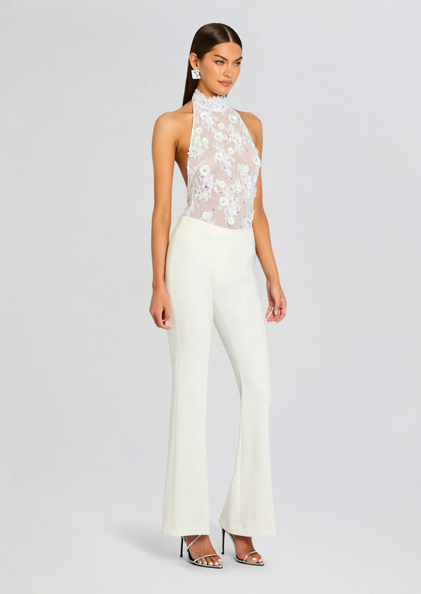 Model wearing a white lace embellished bodysuit with a high neck and sheer floral bodice, paired with ivory trousers.