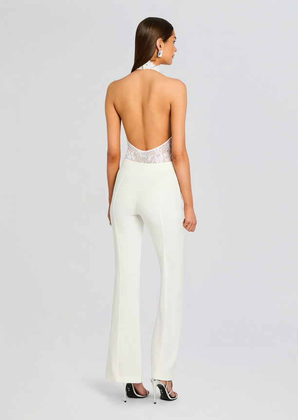 Back view of a model in white lace bodysuit with a low open back and halter neckline, paired with ivory trousers.