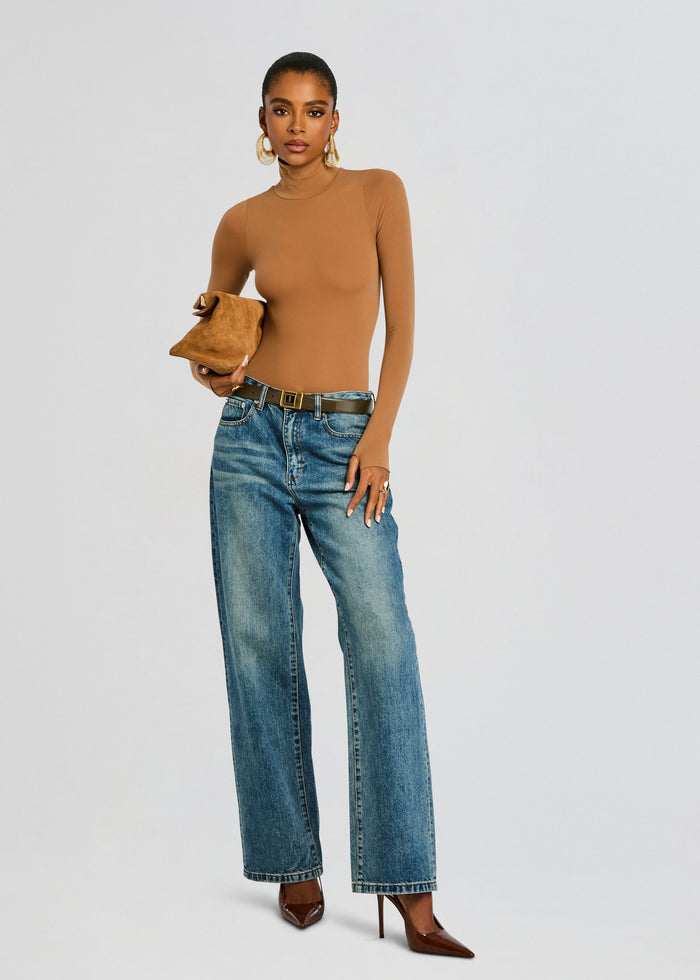 Model wearing camel ballet turtleneck bodysuit with wide-leg jeans, brown belt and brown heels, holding a tan suede clutch. media 2 of 6