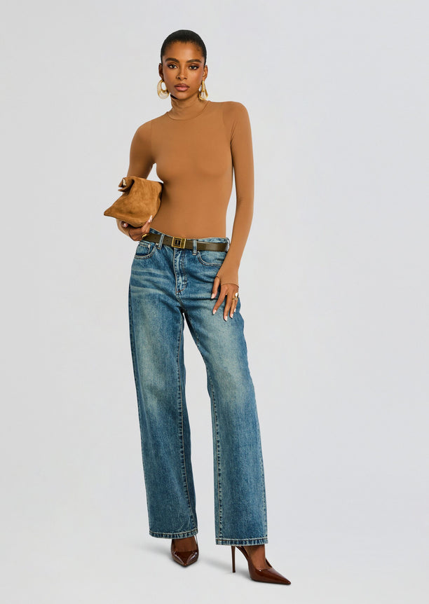 Model wearing camel ballet turtleneck bodysuit with wide-leg jeans, brown belt and brown heels, holding a tan suede clutch.