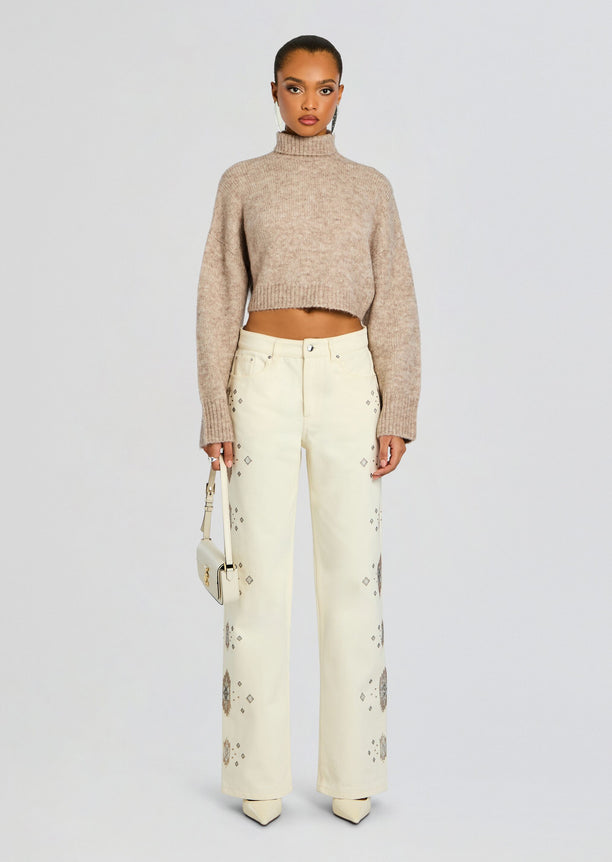 Full body shot of a model in a cropped beige knit sweater and cream embroidered jeans with geometric patterns.
