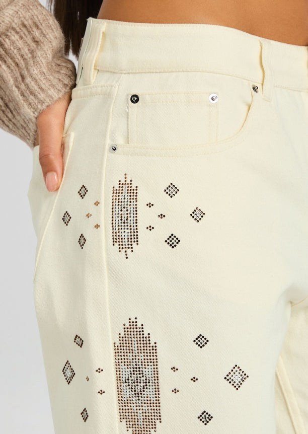 Close-up of pale cream denim at the hip showing a front pocket and geometric rhinestone embroidery along the leg.