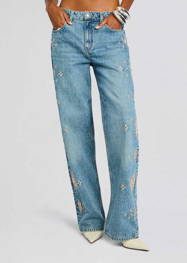 Front view of light-wash Vero Embroidered Jean with pink diamond embroidery down the legs and model with hands in pockets