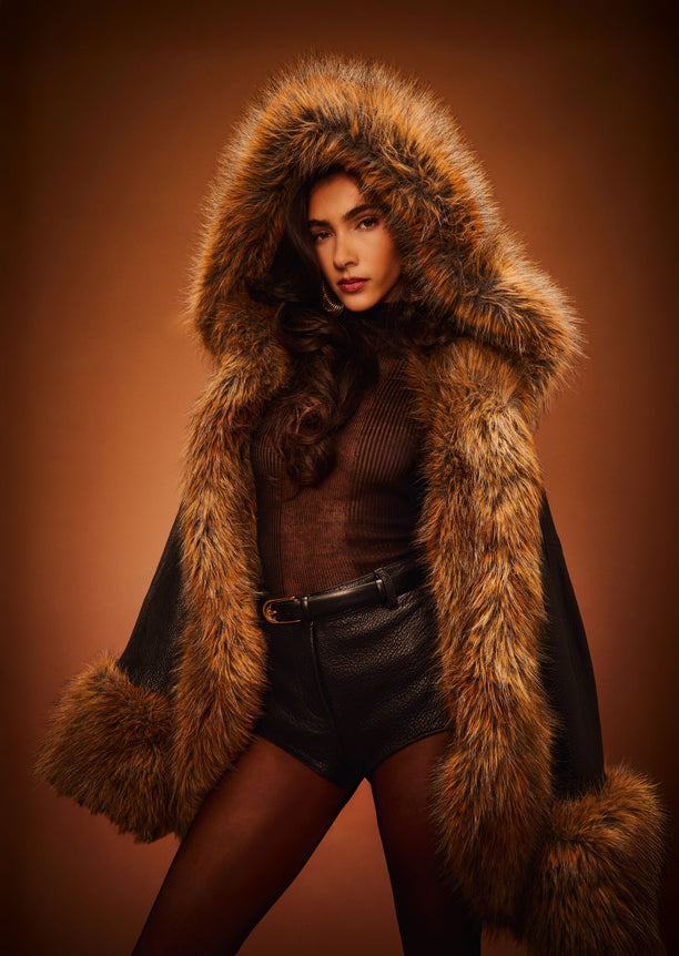Model in a large brown fur hooded coat over a brown knit top and black leather shorts.