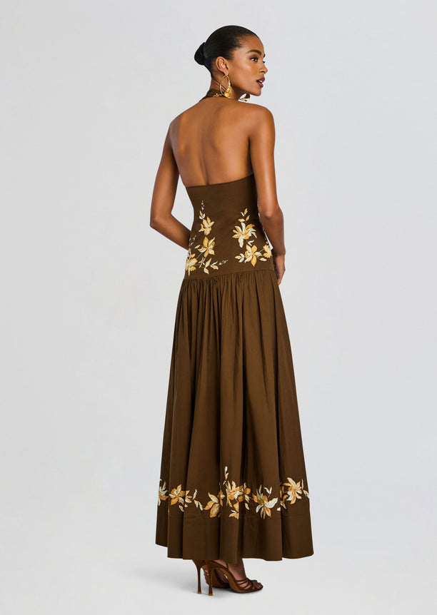 Back view of a brown halter maxi dress with yellow floral embroidery on the bodice and hem, model wearing gold heels.