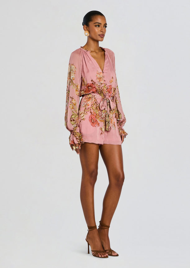 Side view of a model in a pink floral romper with a tied belt and brown strappy heels.