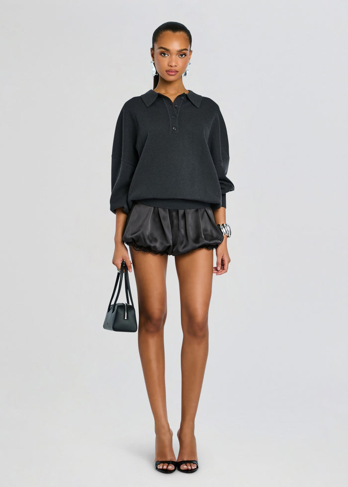 Model in a dark oversized polo sweater and black satin shorts with lace trim, carrying a small black handbag. media 2 of 5