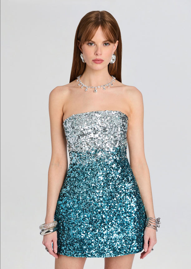 Front view of a strapless sequin mini dress fading from silver at the top to teal at the bottom, model wearing bold jewelry.