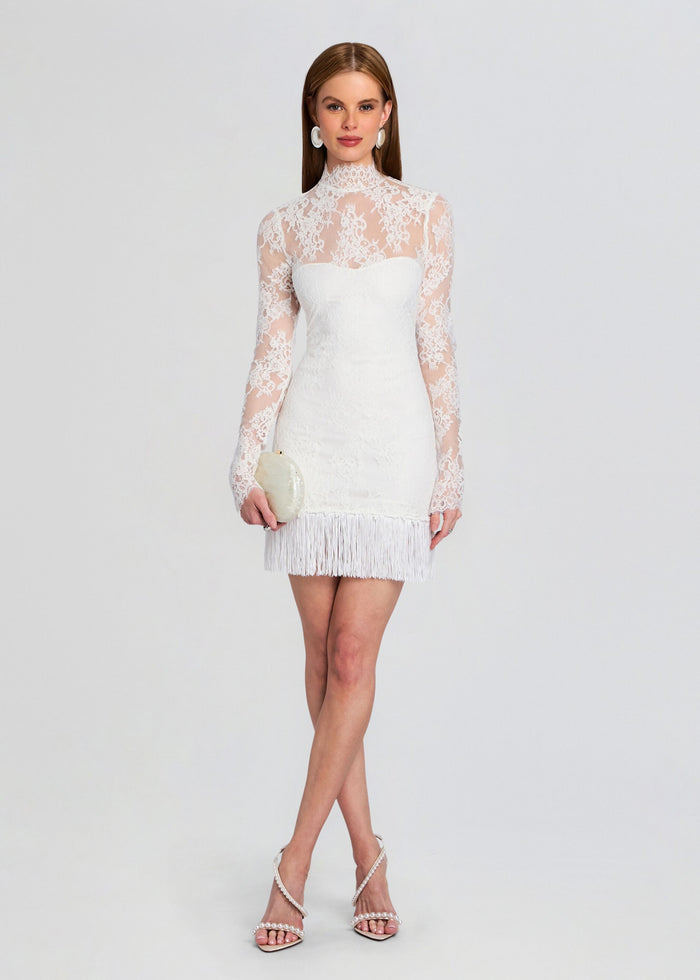 Full length model in a white Xylina Lace Dress with long sleeves and fringe hem, holding a round white clutch. media 1 of 5