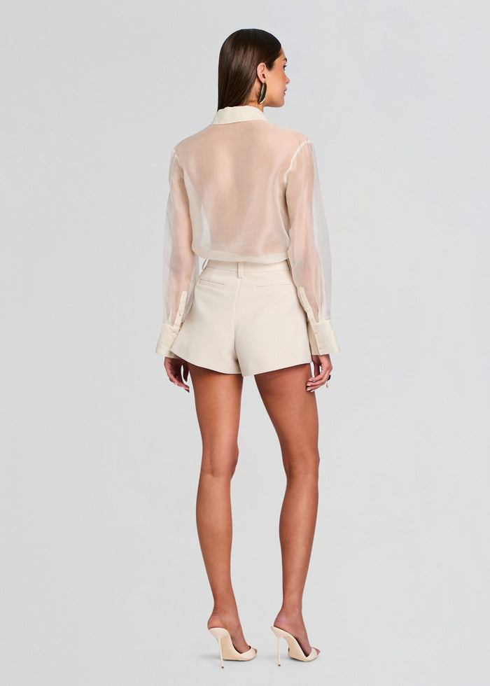 Back view of a model wearing a pale cream sheer blouse tucked into matching high waisted shorts and nude heels media 5 of 5
