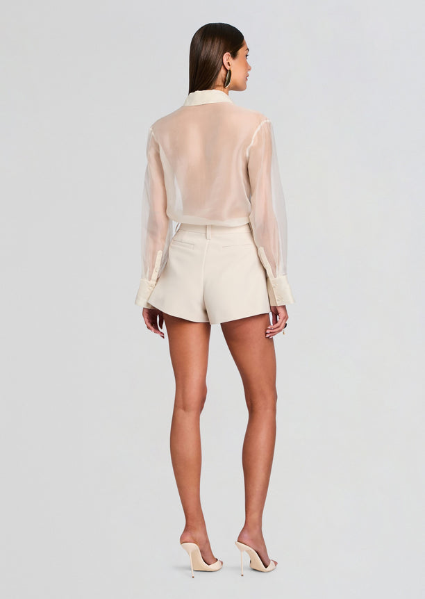 Back view of a model wearing a pale cream sheer blouse tucked into matching high waisted shorts and nude heels