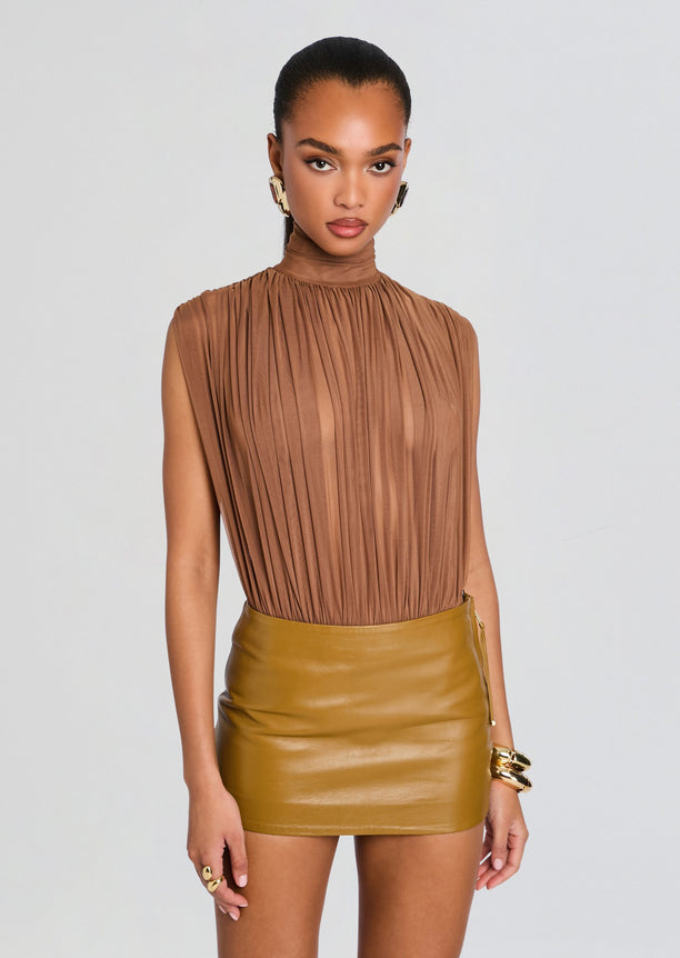 Model wearing a tan pleated high neck sleeveless Giulia Bodysuit paired with a caramel leather mini skirt and gold jewelry.