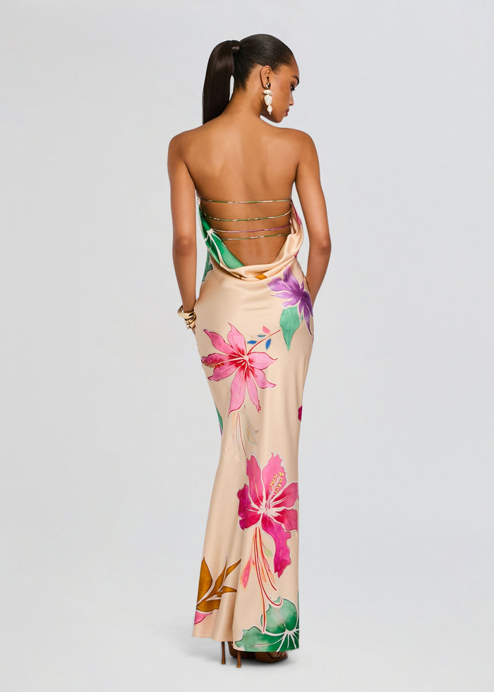 Back view of a model wearing a beige silk dress with large pink hibiscus flowers and green leaves, featuring multiple thin metallic back straps. media 1 of 6