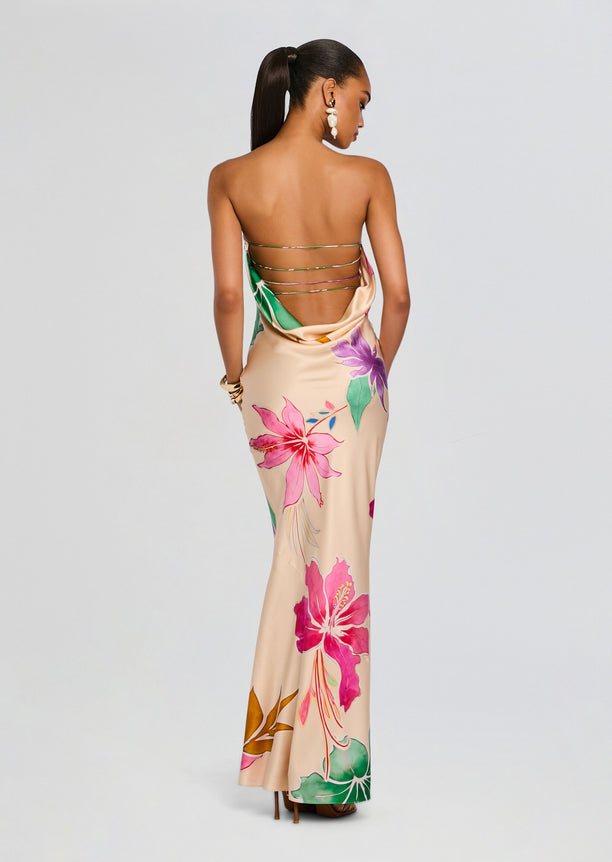 Back view of a model wearing a beige silk dress with large pink hibiscus flowers and green leaves, featuring multiple thin metallic back straps.