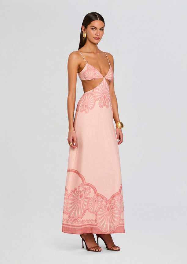 Model wearing Maya Maxi Dress in blush pink with thin straps and waist cutouts, flowing floor-length skirt with a bold decorative hem pattern
