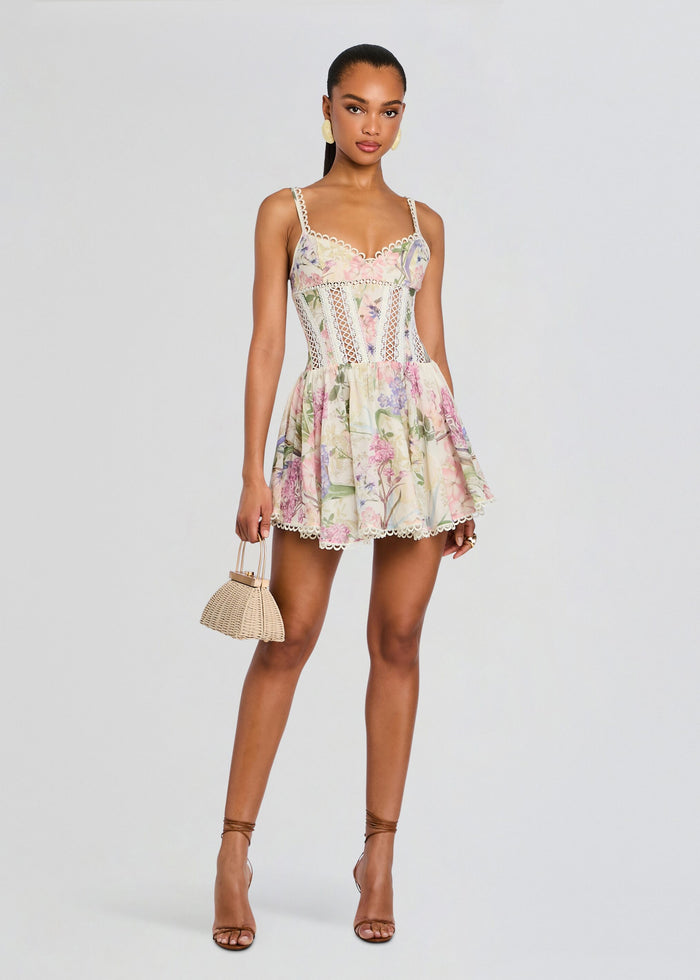Model wearing the Gardenia Short Dress with pastel floral print, lace trim, corset bodice, nude sandals, and a small woven bag media 1 of 5