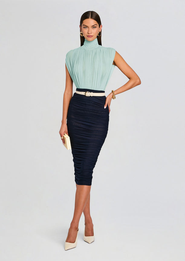 Full-length view of a model in a mint pleated sleeveless high-neck top tucked into a navy ruched midi skirt with a white belt, cream heels, and a small clutch.