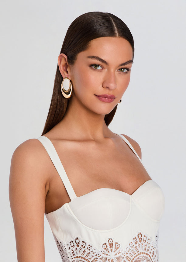 Model wearing oversized gold-tone hoop earring with white oval centerpiece.