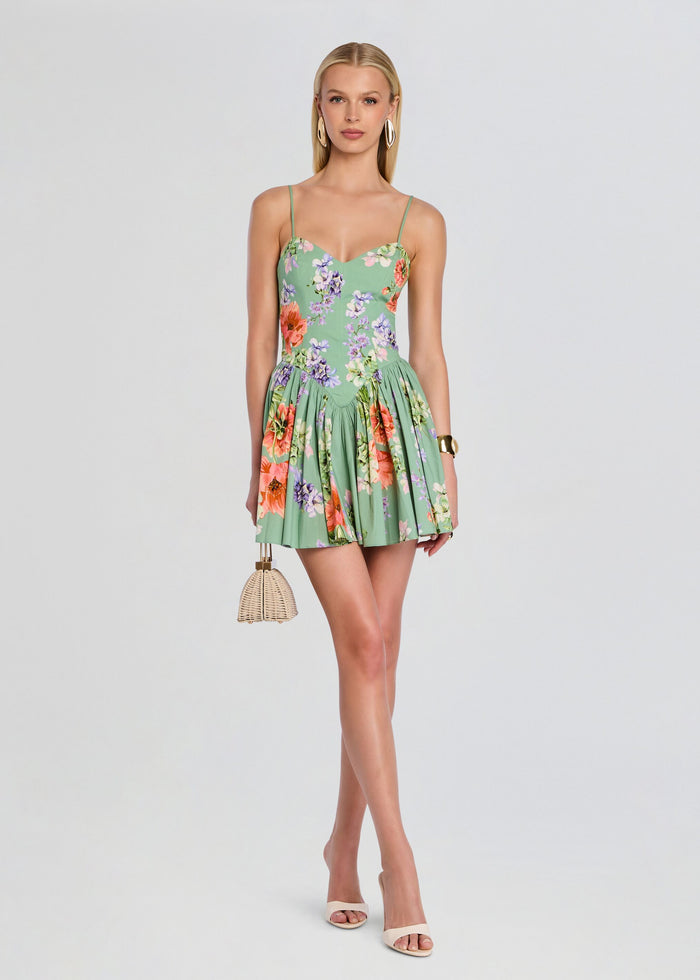 Full body shot of a model in a green floral Cove Corseted Mini Dress with thin straps and white sandals, carrying a wicker bag. media 1 of 5