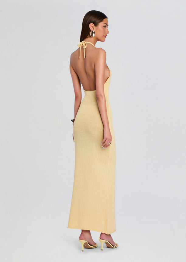 Back view of a model in a pale yellow knit halter maxi dress showing an open back and neck tie.