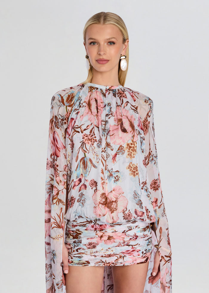 Model wearing Ezra Ruched Cape Mini Dress in a pale floral print with sheer long sleeves and a ruched mini skirt media 5 of 5
