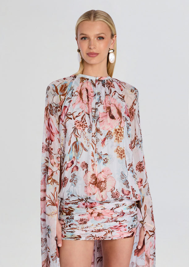 Model wearing Ezra Ruched Cape Mini Dress in a pale floral print with sheer long sleeves and a ruched mini skirt